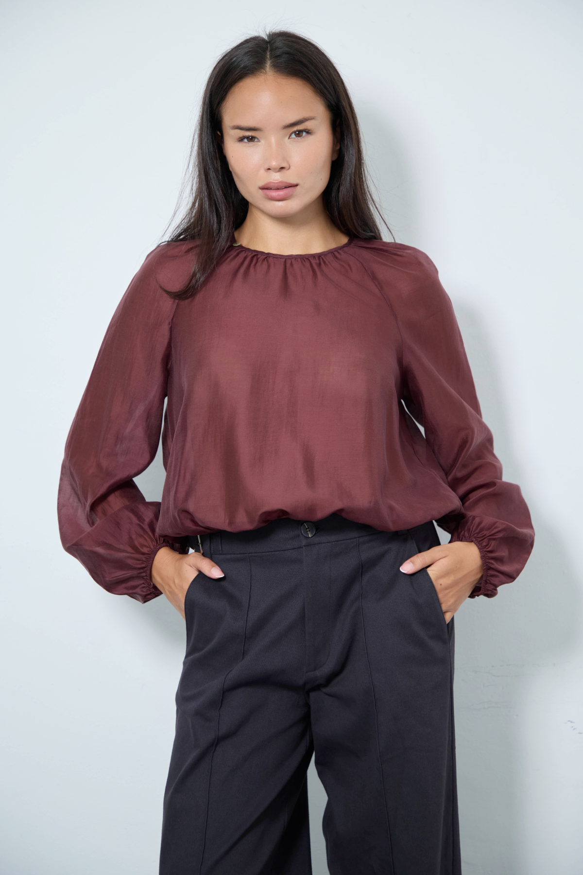 A woman modeling a long-sleeve blouse with a gathered, balloon-style sleeve and a round neckline in a deep burgundy color, paired with dark charcoal trousers, against a light gray studio background.